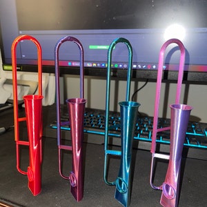 May include: Four colorful 3D printed metal pipes, each with a different color gradient. The pipes are arranged in a row on a dark surface. The colors are red, purple, teal, and pink.