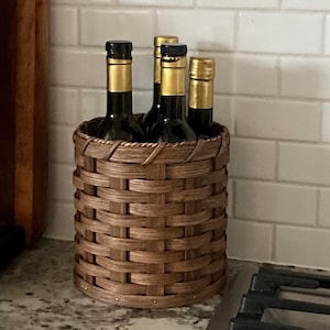 May include: A brown wicker basket holds five glass bottles with gold tops. The bottles are arranged in a circle and are all the same size. The basket is sitting on a countertop in a kitchen.