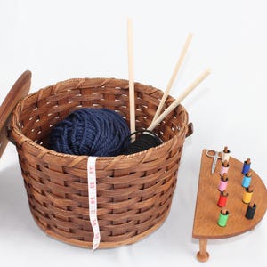 Puede incluir: Una cesta de mimbre marr&oacute;n con tapa, que contiene una bola de hilo azul y agujas de tejer de madera. Un soporte de madera con carretes de hilo de colores y un par de tijeras est&aacute; al lado de la cesta.