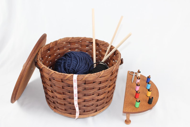 Puede incluir: Una cesta de mimbre marr&oacute;n con tapa, que contiene una bola de hilo azul y agujas de tejer de madera. Un soporte de madera con carretes de hilo de colores y un par de tijeras est&aacute; al lado de la cesta.