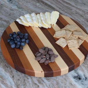 Puede incluir: Una tabla de servir redonda de madera con rayas alternas de madera clara y oscura. La tabla est&aacute; llena de aperitivos, incluyendo una cu&ntilde;a de queso brie, ar&aacute;ndanos, almendras y galletas.
