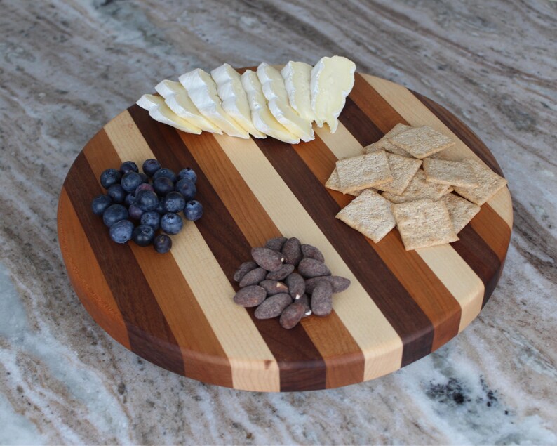 Puede incluir: Una tabla de servir redonda de madera con rayas alternas de madera clara y oscura. La tabla est&aacute; llena de aperitivos, incluyendo una cu&ntilde;a de queso brie, ar&aacute;ndanos, almendras y galletas.