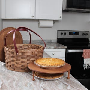 Puede incluir: Una cesta de mimbre marr&oacute;n con asas rojas y una tapa de madera. Dos tartas est&aacute;n en un soporte de madera de dos niveles delante de la cesta. Las tartas est&aacute;n sobre una encimera de cocina blanca.