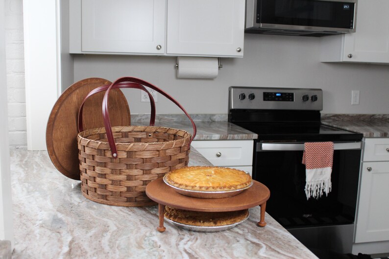 Puede incluir: Una cesta de mimbre marr&oacute;n con asas rojas y una tapa de madera. Dos tartas est&aacute;n en un soporte de madera de dos niveles delante de la cesta. Las tartas est&aacute;n sobre una encimera de cocina blanca.