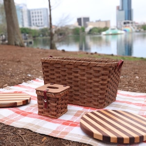 Puede incluir: Un conjunto de cesta de picnic con una cesta tejida, una caja tejida m&aacute;s peque&ntilde;a y dos tablas de cortar redondas de madera con patrones rayados. El conjunto est&aacute; sobre una manta de picnic a cuadros rojos y blancos.