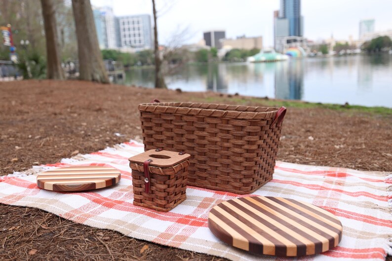 Puede incluir: Un conjunto de cesta de picnic con una cesta tejida, una caja tejida m&aacute;s peque&ntilde;a y dos tablas de cortar redondas de madera con patrones rayados. El conjunto est&aacute; sobre una manta de picnic a cuadros rojos y blancos.