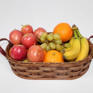 Puede incluir: Una cesta de mimbre tejida con asas de cuero llena de fruta. La cesta contiene manzanas rojas, uvas verdes, pl&aacute;tanos amarillos y naranjas.