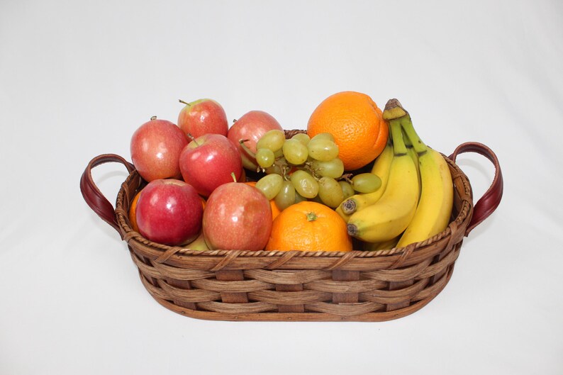 Puede incluir: Una cesta de mimbre tejida con asas de cuero llena de fruta. La cesta contiene manzanas rojas, uvas verdes, pl&aacute;tanos amarillos y naranjas.