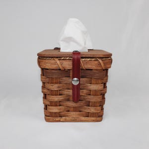 Puede incluir: Un cubre caja de pa&ntilde;uelos de mimbre marr&oacute;n con una correa de cuero roja y un bot&oacute;n plateado. La tapa es de forma cuadrada y est&aacute; hecha de mimbre tejido.