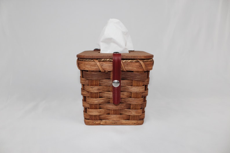 Puede incluir: Un cubre caja de pa&ntilde;uelos de mimbre marr&oacute;n con una correa de cuero roja y un bot&oacute;n plateado. La tapa es de forma cuadrada y est&aacute; hecha de mimbre tejido.