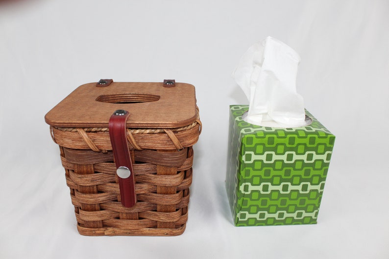 K&ouml;nnte beinhalten: Zwei Taschentuchboxen. Eine ist ein geflochtener Weidenkorb mit einem Holzdeckel und einem Lederriemen. Die andere ist eine Schachtel aus Pappe mit einem gr&uuml;n-wei&szlig;en geometrischen Muster.