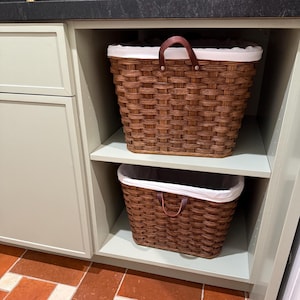 May include: Two woven brown baskets with white liners and brown leather handles are stacked on a shelf. The baskets are inside a light green cabinet. The baskets are designed for storage.
