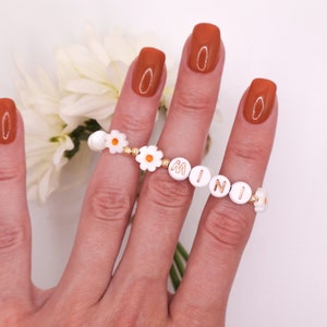 May include: A white daisy charm bracelet with gold beads and the word "MINI" spelled out in gold letters. The bracelet is worn on a hand with orange nail polish.