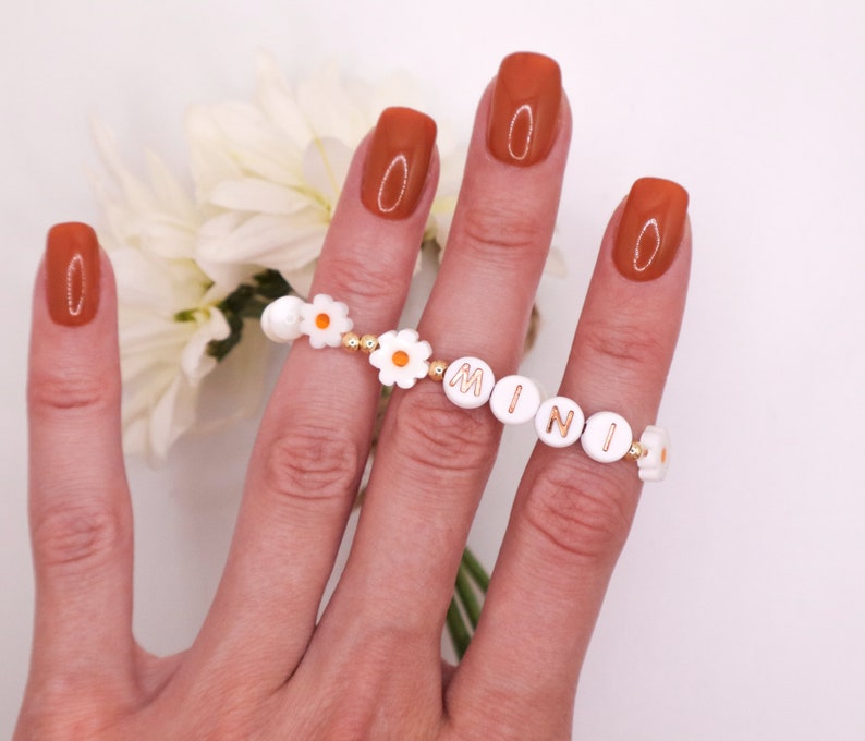 May include: A white daisy charm bracelet with gold beads and the word "MINI" spelled out in gold letters. The bracelet is worn on a hand with orange nail polish.