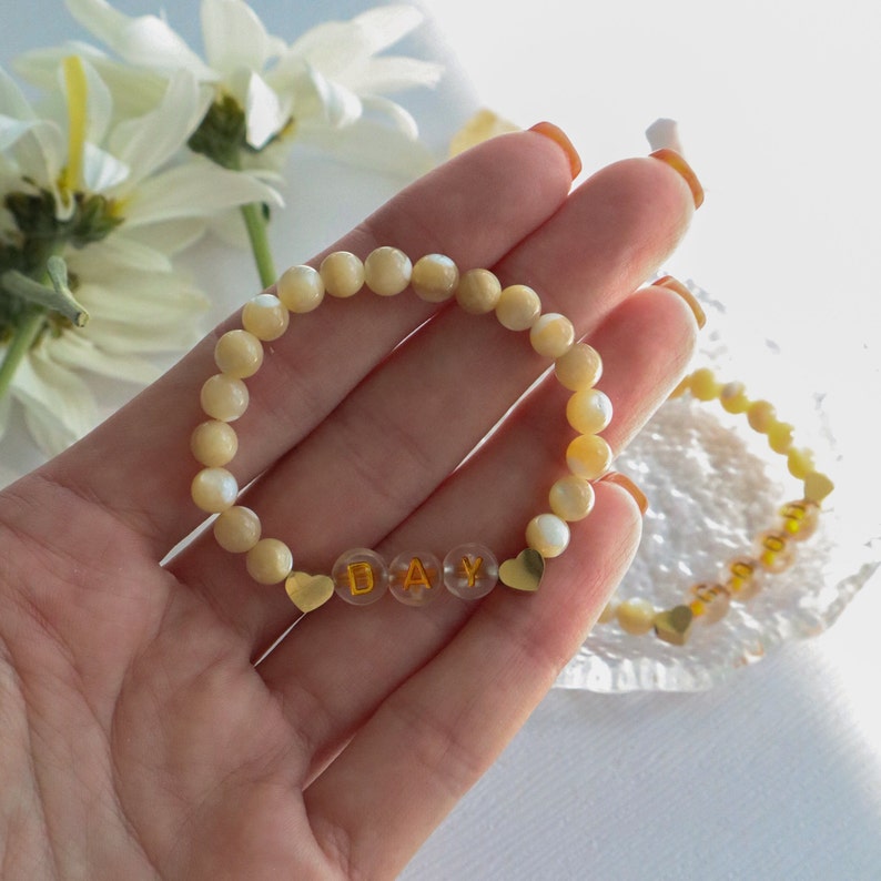 May include: A white beaded bracelet with gold letter beads spelling "DAY" and gold heart charms on either side.