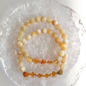 May include: Two bracelets with gold letter beads spelling "GOOD DAY" on a white background. The beads are made of mother of pearl and have a light beige color.