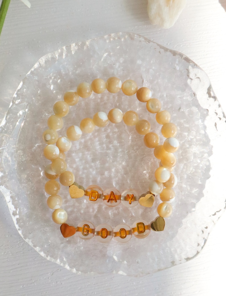 May include: Two bracelets with gold letter beads spelling "GOOD DAY" on a white background. The beads are made of mother of pearl and have a light beige color.