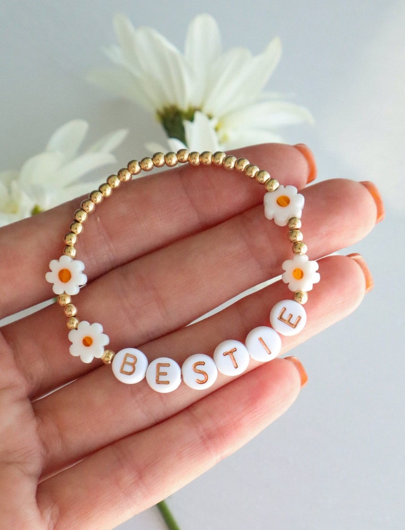 May include: A gold beaded bracelet with white daisy beads and gold letter beads spelling out "BESTIE".