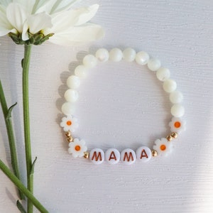 May include: White beaded bracelet with white daisy beads and gold accents. The bracelet spells out "MAMA" in gold letters.