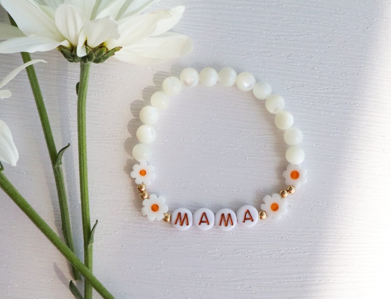 May include: White beaded bracelet with white daisy beads and gold accents. The bracelet spells out "MAMA" in gold letters.