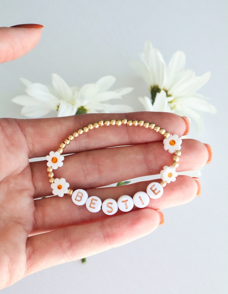 May include: A gold beaded bracelet with white daisy beads and white letter beads spelling "BESTIE".