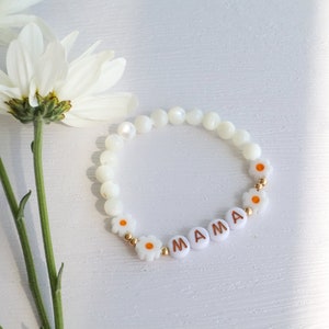 May include: White beaded bracelet with gold accents and white daisy charms spelling out "MAMA".