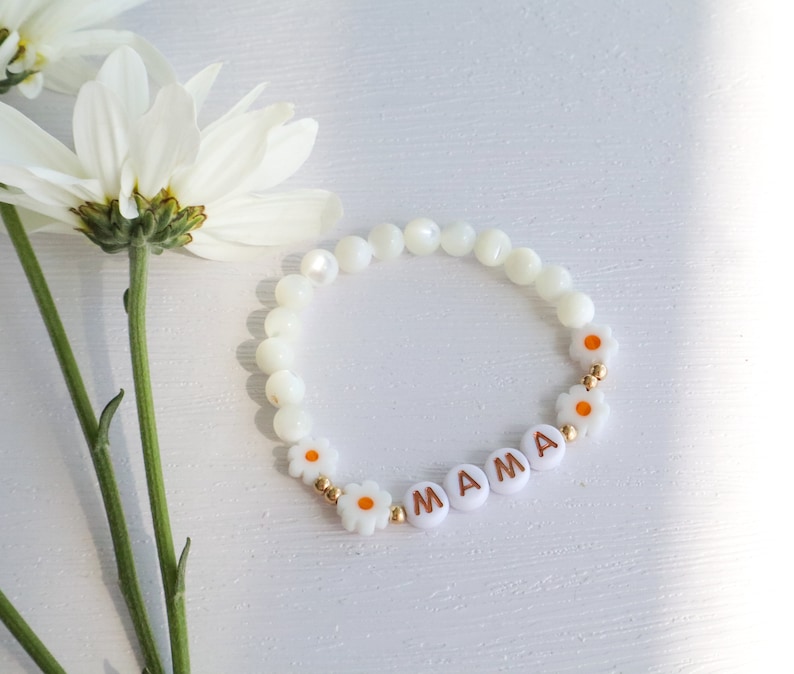 May include: White beaded bracelet with gold accents and white daisy charms spelling out "MAMA".