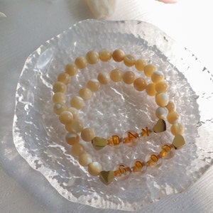 May include: Two beaded bracelets with gold heart charms and letter beads spelling "GOOD DAY". The bracelets are on a clear glass dish.