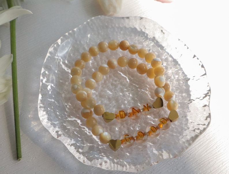 May include: Two beaded bracelets with gold heart charms and letter beads spelling "GOOD DAY". The bracelets are on a clear glass dish.