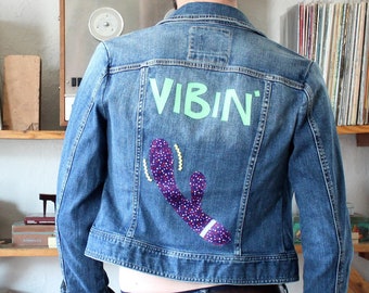 May include: A blue denim jacket featuring the word VIBIN in light green letters on the back, accompanied by a purple graphic of a curved, dotted object with yellow wavy lines. The jacket has a classic collar, long sleeves, and buttoned cuffs.