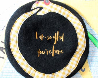 May include: A black felt beret featuring an embroidered yellow and white gingham snake forming a circle. The snake has two small red googly eyes. Centered inside the snake is the gold embroidered text: I am so glad you're here.