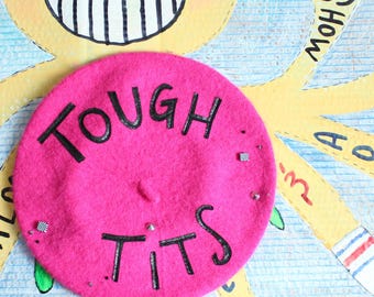 May include: A bright pink felt beret featuring the black embroidered text "TOUGH TITS" across the center. The beret is decorated with small silver-toned metal studs and square accents, positioned over a yellow illustrated character background.