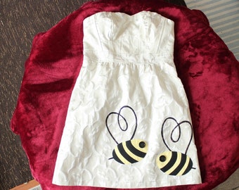 May include: A strapless white dress featuring a subtle tonal floral jacquard pattern and a fitted bodice. The lower portion of the dress is decorated with two appliqued bumblebee designs, each featuring yellow and black striped bodies.