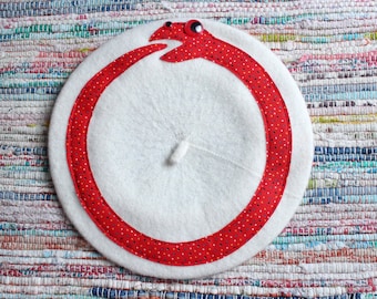 May include: A white felt beret featuring a red snake appliqué with small multicolored dots. The snake is positioned in a circular Ouroboros shape, biting its own tail, and includes a prominent black and white eye detail on the head.