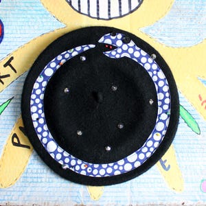 May include: A black beret featuring a circular applique of a blue and white polka-dot snake biting its own tail. The snake design is accented with small black buttons for eyes, tiny red beads for a tongue, and several silver-toned metal studs.
