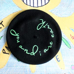 May include: A black beret featuring light green embroidered text that reads "you and i are earth" in a circular pattern around the center. The embroidery is stitched in a cursive script style, contrasting against the solid black fabric surface.