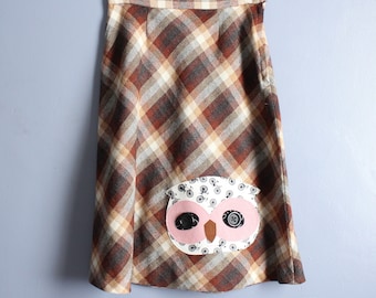 May include: A brown, tan, and red plaid skirt with an owl applique. The owl has pink and brown accents, with black button eyes and a bicycle print on the wings. The skirt is hanging on a clear hanger.