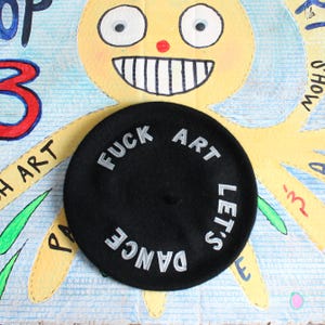 May include: A black beret featuring the embroidered silver text "FUCK ART LET'S DANCE" arranged in a circular pattern around the center. The hat has a smooth, felt-like texture and a small, raised knot at the very top of the crown.