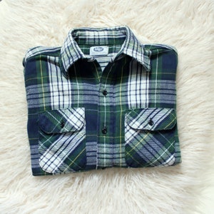 May include: A long-sleeve button-down shirt featuring a plaid pattern in shades of dark green, navy blue, white, and thin yellow lines. The shirt includes a pointed collar, a front button placket, and two chest pockets with buttoned flaps.
