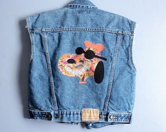 May include: A sleeveless denim vest in light blue with a cartoon graphic on the back. The graphic features a smiling character wearing sunglasses and a beret. The vest has a "French Toast" label.