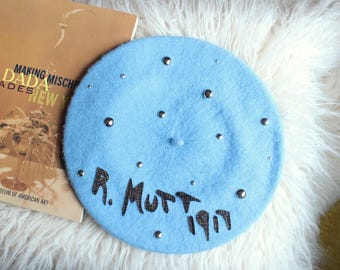 May include: A light blue beret featuring scattered silver-toned studs and the text R. MUTT 1917 in a dark, textured, grid-patterned font. The beret has a small central knob and a soft, fuzzy texture, presented as a flat circular accessory.