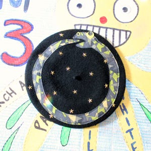 May include: A black beret featuring a circular graphic of a snake with a green and grey camouflage pattern. The snake is depicted biting its own tail, and the design is accented with small, metallic gold-colored star-shaped studs.