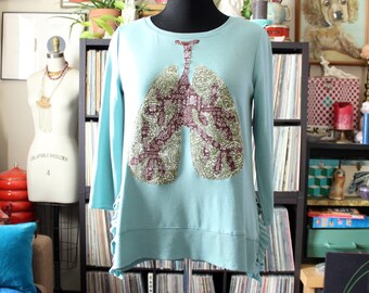 May include: A light blue long-sleeved shirt with a unique design of lungs. The lungs are patterned with a mix of olive green and burgundy paisley fabric. The shirt has a crew neck and decorative side ties.