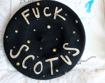 May include: A black beret featuring the text FUCK SCOTUS in a grid-patterned fabric appliqué. The hat is decorated with scattered gold and dark metallic studs across the surface, creating a textured appearance on the dark felt material.