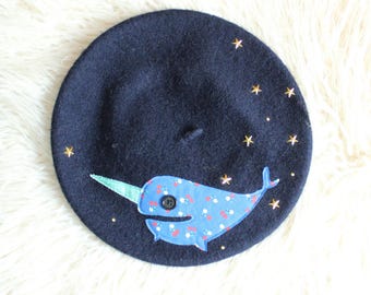 May include: A dark navy blue wool beret featuring an appliqued blue narwhal with a floral pattern and a black button eye. The narwhal has a light green horn, and the beret is decorated with several small gold-toned metal star-shaped studs.