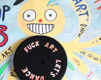 May include: A black beret featuring the text FUCK ART LET'S DANCE in pink lettering. The beret is positioned over a yellow illustrated character with a wide grin, googly eyes, and a black beret on its head, set against a blue background.