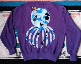 May include: A purple long-sleeve sweater featuring a large octopus appliqué on the back. The octopus is constructed from white, blue, and black patterned fabric with a small yellow crown above its head and two dark circular buttons for eyes.