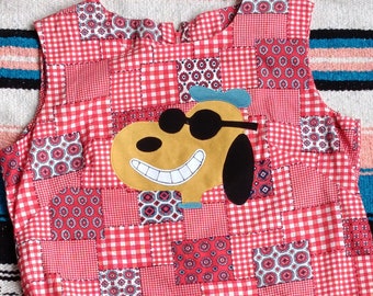 May include: A sleeveless dress featuring a patchwork pattern of red and white gingham and floral geometric prints. The center displays a large applique of a tan dog wearing black sunglasses and a blue beret, with a wide, toothy white grin.
