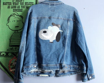 May include: A medium-wash denim jacket featuring a cartoon character on the back. The character is white, black, and light blue, with a smiling expression. The jacket has a classic design with button closures and pockets. A green banner with the text "IT DOESN'T MATTER WHAT YOU BELIEVE AS LONG AS YOU'RE SINCERE!" hangs in the background.
