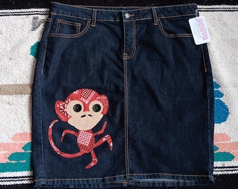 May include: A dark blue denim skirt featuring a red and white patterned monkey appliqué on the lower left side. The monkey has two black button eyes. The skirt has a standard waistband with a metal button, front pockets, and a white brand tag.